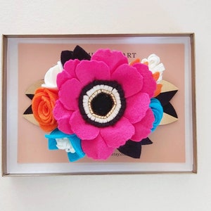 May include: A pink felt flower with a black center and white and gold accents, surrounded by smaller felt flowers in orange, white, blue, and black. The flowers are arranged on a wooden base with a black felt ribbon.