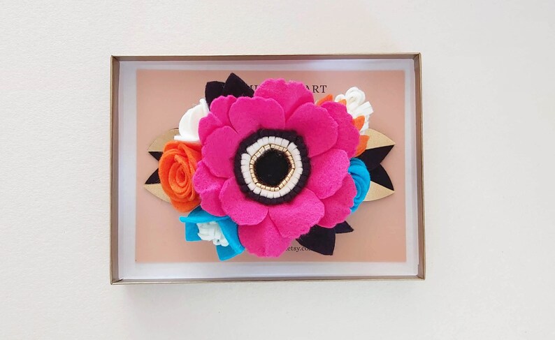 May include: A pink felt flower with a black center and white and gold accents, surrounded by smaller felt flowers in orange, white, blue, and black. The flowers are arranged on a wooden base with a black felt ribbon.
