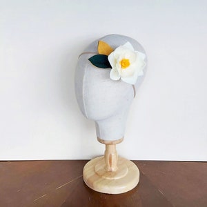 May include: A white felt flower headband with a yellow center and green leaves. The headband is displayed on a white mannequin head.