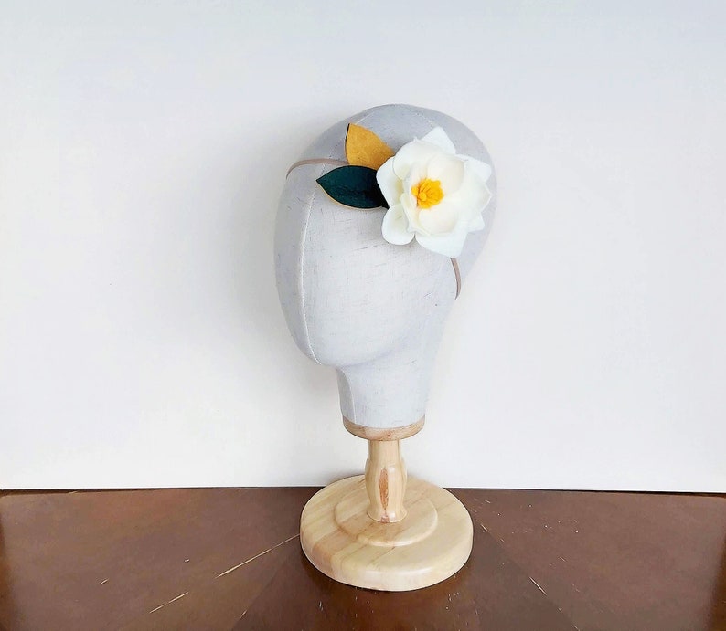May include: A white felt flower headband with a yellow center and green leaves. The headband is displayed on a white mannequin head.
