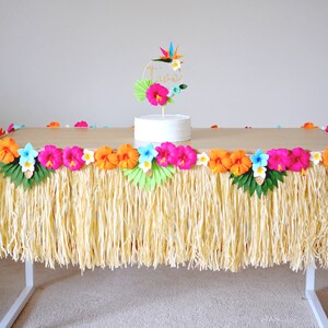 May include: A table decorated with a grass skirt and colourful paper flowers. The table is adorned with orange, pink, and blue hibiscus flowers, and a cake with a floral topper. The table is set for a luau or tropical-themed party.