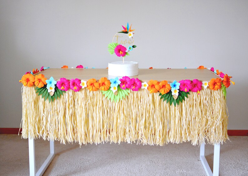 May include: A table decorated with a grass skirt and colourful paper flowers. The table is adorned with orange, pink, and blue hibiscus flowers, and a cake with a floral topper. The table is set for a luau or tropical-themed party.