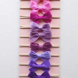 May include: A set of eight felt hair bows in shades of pink, purple, and lavender, attached to nude elastic headbands. The bows are arranged in a gradient from light pink to dark purple. The headbands are displayed on a pink background with the text "etsy.com" at the bottom.