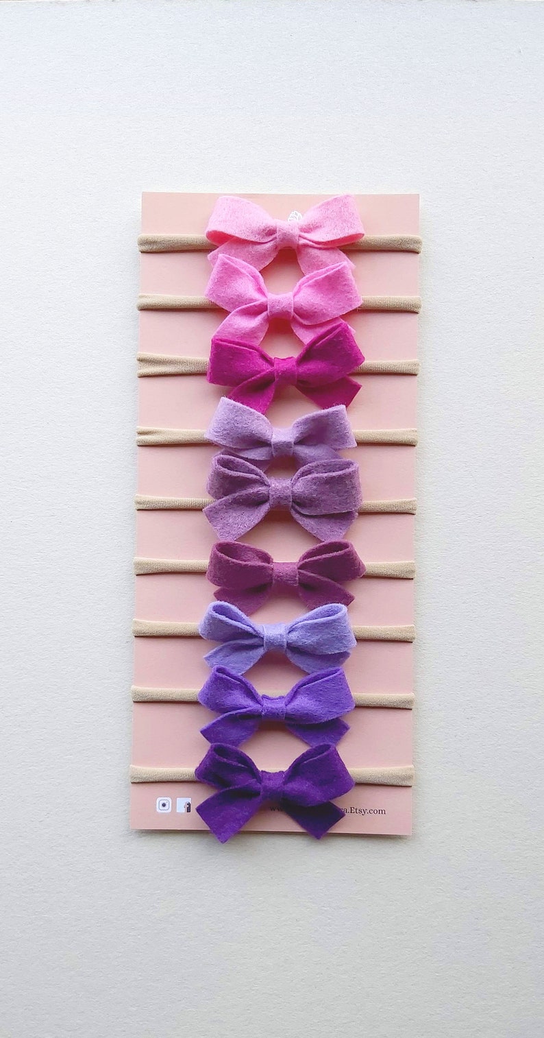 May include: A set of eight felt hair bows in shades of pink, purple, and lavender, attached to nude elastic headbands. The bows are arranged in a gradient from light pink to dark purple. The headbands are displayed on a pink background with the text "etsy.com" at the bottom.