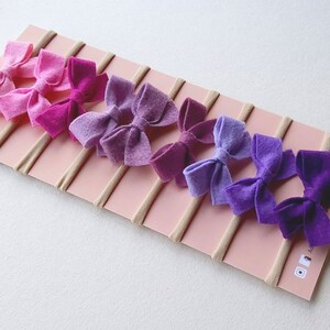 May include: Set of eight felt hair bows in shades of purple and pink, attached to nude elastic headbands. The bows are arranged in a gradient from light pink to dark purple.