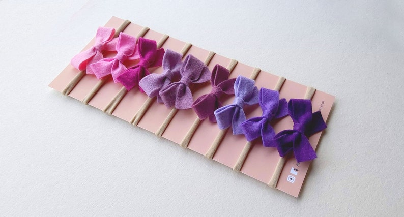 May include: Set of eight felt hair bows in shades of purple and pink, attached to nude elastic headbands. The bows are arranged in a gradient from light pink to dark purple.