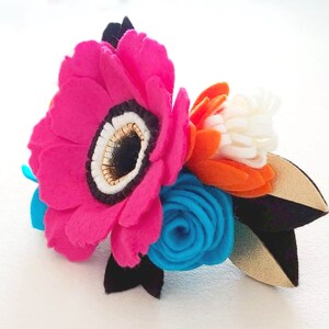 May include: A felt flower bouquet with a large pink flower, a smaller blue flower, and an orange flower with white accents. The flowers are arranged on a black and gold felt stem.
