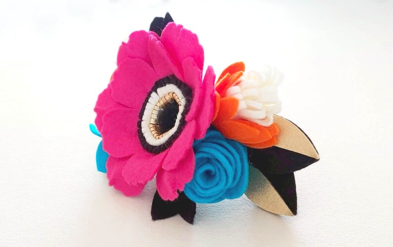 May include: A felt flower bouquet with a large pink flower, a smaller blue flower, and an orange flower with white accents. The flowers are arranged on a black and gold felt stem.