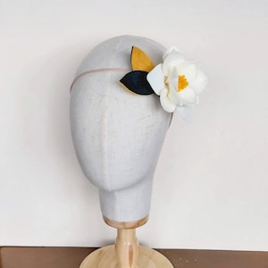 May include: A white felt flower with yellow center and green leaves attached to a headband. The headband is on a white mannequin head.