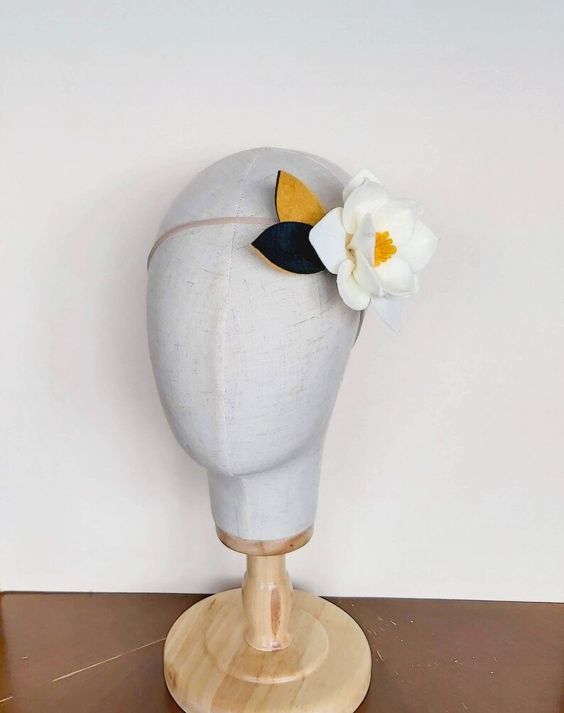 May include: A white felt flower with yellow center and green leaves attached to a headband. The headband is on a white mannequin head.