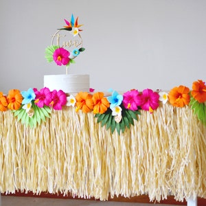 May include: A party table decorated with a grass skirt, colourful paper flowers, and a cake with a gold "two" topper. The flowers are pink, orange, and blue, creating a tropical theme for a birthday celebration.