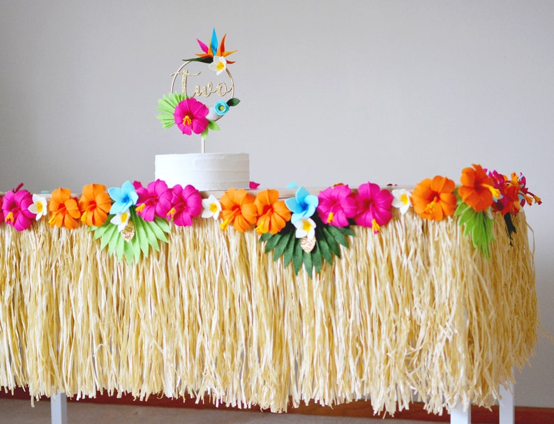 May include: A party table decorated with a grass skirt, colourful paper flowers, and a cake with a gold "two" topper. The flowers are pink, orange, and blue, creating a tropical theme for a birthday celebration.