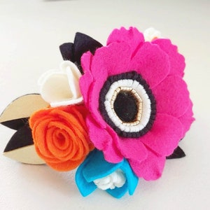 May include: A felt flower arrangement with a large pink flower, a smaller orange rose, a white flower, and a blue flower. The flowers are attached to a wooden base.