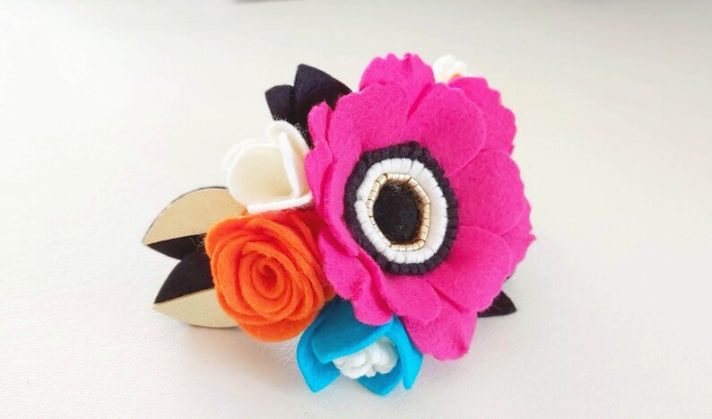 May include: A felt flower arrangement with a large pink flower, a smaller orange rose, a white flower, and a blue flower. The flowers are attached to a wooden base.