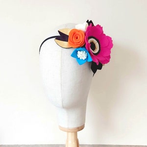 May include: A black headband with a felt flower arrangement. The flowers are pink, orange, white, and blue. The headband is on a white mannequin head.