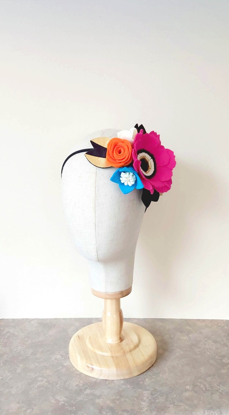 May include: A black headband with a felt flower arrangement. The flowers are pink, orange, white, and blue. The headband is on a white mannequin head.