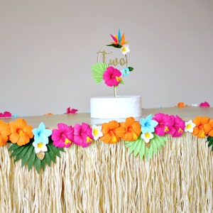 May include: A tropical-themed party table setting. The table is adorned with a grass skirt and a garland of colourful paper flowers. A cake topper with the word "two" is also present. The flowers are in shades of pink, orange, and blue.