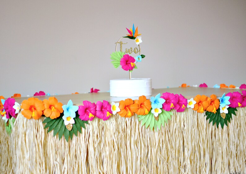 May include: A tropical-themed party table setting. The table is adorned with a grass skirt and a garland of colourful paper flowers. A cake topper with the word "two" is also present. The flowers are in shades of pink, orange, and blue.