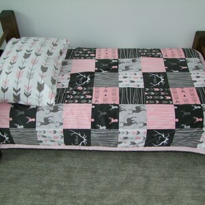May include: A small wooden bed frame with a patchwork quilt and pillow. The quilt features pink, gray, and black squares with deer, arrows, and text. The pillow has a white background with gray and pink arrows.