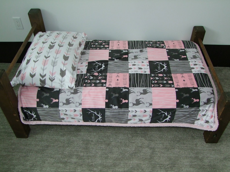 May include: A small wooden bed frame with a patchwork quilt and pillow. The quilt features pink, gray, and black squares with deer, arrows, and text. The pillow has a white background with gray and pink arrows.