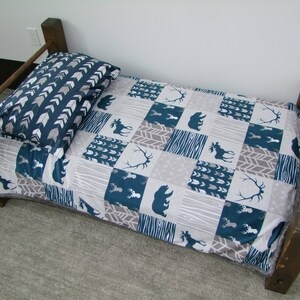 May include: A small wooden bed frame with a patterned quilt and pillow. The quilt features squares with bear, moose, and antler designs in teal and gray. The pillow has a teal and white arrow pattern.