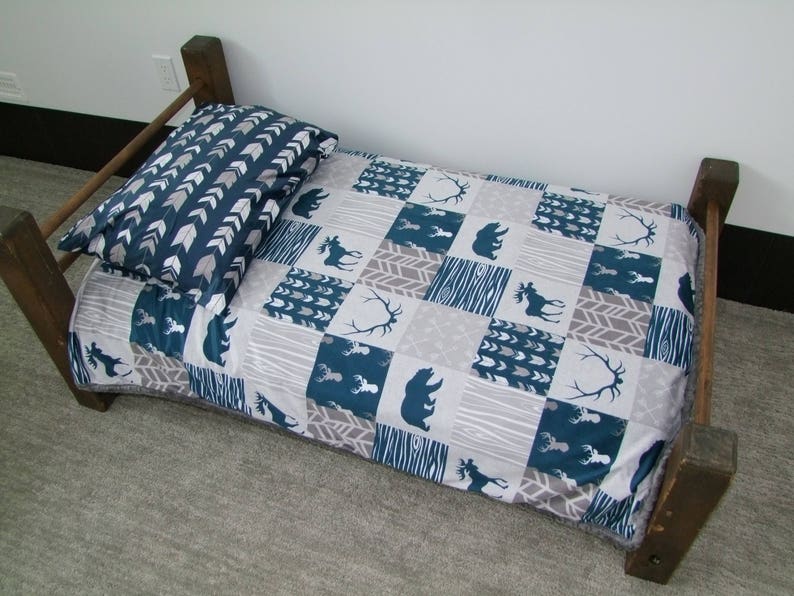 May include: A small wooden bed frame with a patterned quilt and pillow. The quilt features squares with bear, moose, and antler designs in teal and gray. The pillow has a teal and white arrow pattern.