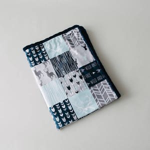 May include: A patchwork baby blanket with a navy blue border. The blanket features various patterns, including deer, arrows, and geometric shapes in shades of blue, gray, and white. The text "WILD AND FREE" is also visible.