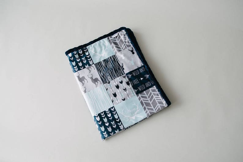 May include: A patchwork baby blanket with a navy blue border. The blanket features various patterns, including deer, arrows, and geometric shapes in shades of blue, gray, and white. The text "WILD AND FREE" is also visible.