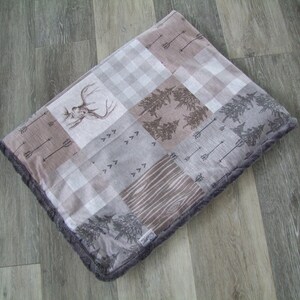 May include: A square, patchwork-style pet bed with a woodland theme. The bed features various patterned squares, including a deer head, arrows, trees, and wood grain. The edges are trimmed with a dark gray, plush material.