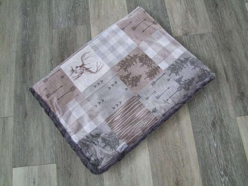 May include: A square, patchwork-style pet bed with a woodland theme. The bed features various patterned squares, including a deer head, arrows, trees, and wood grain. The edges are trimmed with a dark gray, plush material.