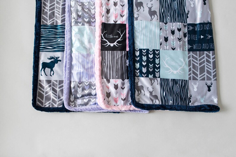 May include: A collection of patchwork baby blankets with various patterns and colors. Designs include arrows, deer, and wood grain. The blankets have a soft, plush texture and are trimmed with a dark blue border. One blanket has the text "little one".