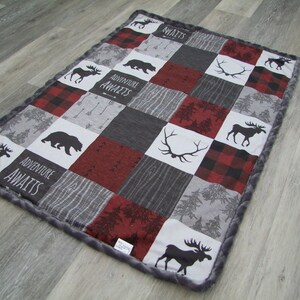 May include: A patchwork baby blanket with a gray, red, and black color scheme. The blanket features squares with moose, bear, and antler silhouettes, along with the words "ADVENTURE AWAITS." The border is a soft gray material.