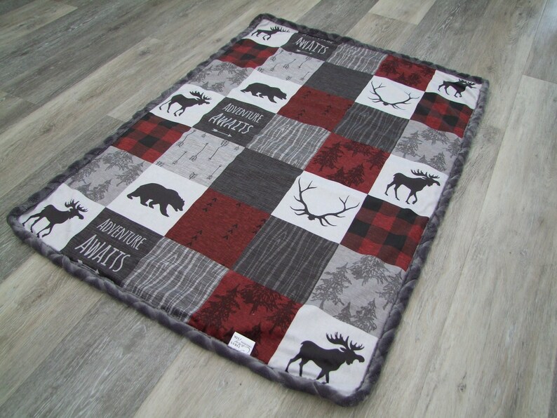 May include: A patchwork baby blanket with a gray, red, and black color scheme. The blanket features squares with moose, bear, and antler silhouettes, along with the words "ADVENTURE AWAITS." The border is a soft gray material.