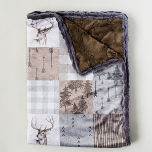 May include: A patchwork baby blanket with a deer, arrow, tree, and plaid pattern. The blanket has a brown, plush interior and a gray, textured border. The exterior features various neutral-toned squares with nature-themed designs.