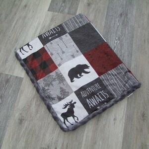 May include: A patchwork blanket featuring a bear, moose, and arrow design. The blanket has a gray, red, and black color scheme with the words "Adventure Awaits" and "Awaits" printed on it. The blanket has a gray, plush border.