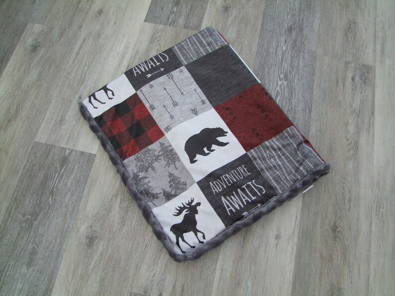May include: A patchwork blanket featuring a bear, moose, and arrow design. The blanket has a gray, red, and black color scheme with the words "Adventure Awaits" and "Awaits" printed on it. The blanket has a gray, plush border.