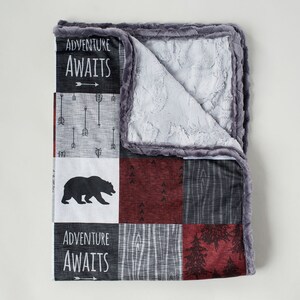 May include: A patchwork baby blanket with a gray, red, and black color scheme. The blanket features a bear silhouette, arrows, trees, and the text "ADVENTURE AWAITS". The border is a soft gray faux fur.
