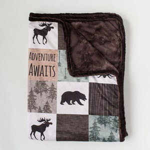 Peut inclure: Une couverture patchwork avec une bordure en fausse fourrure marron. La couverture présente des carrés avec des motifs sur le thème de la forêt, notamment des élans, des ours, des arbres et le texte "ADVENTURE AWAITS". La palette de couleurs comprend le marron, le gris et le blanc.