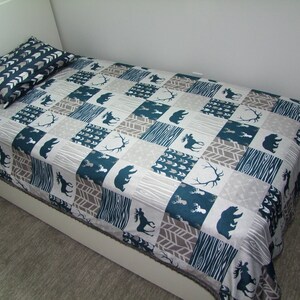 May include: A twin-sized bed with a matching pillow and a patchwork quilt. The quilt features a pattern of teal, gray, and white squares with bear, moose, and antler silhouettes. The pillow has a chevron pattern.