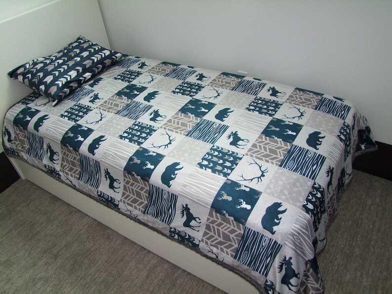 May include: A twin-sized bed with a matching pillow and a patchwork quilt. The quilt features a pattern of teal, gray, and white squares with bear, moose, and antler silhouettes. The pillow has a chevron pattern.