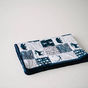 May include: A folded patchwork blanket featuring a variety of patterns and animal silhouettes in teal and gray on a white background. The blanket has a plush, dark blue border. The design includes bears, moose, and abstract patterns.