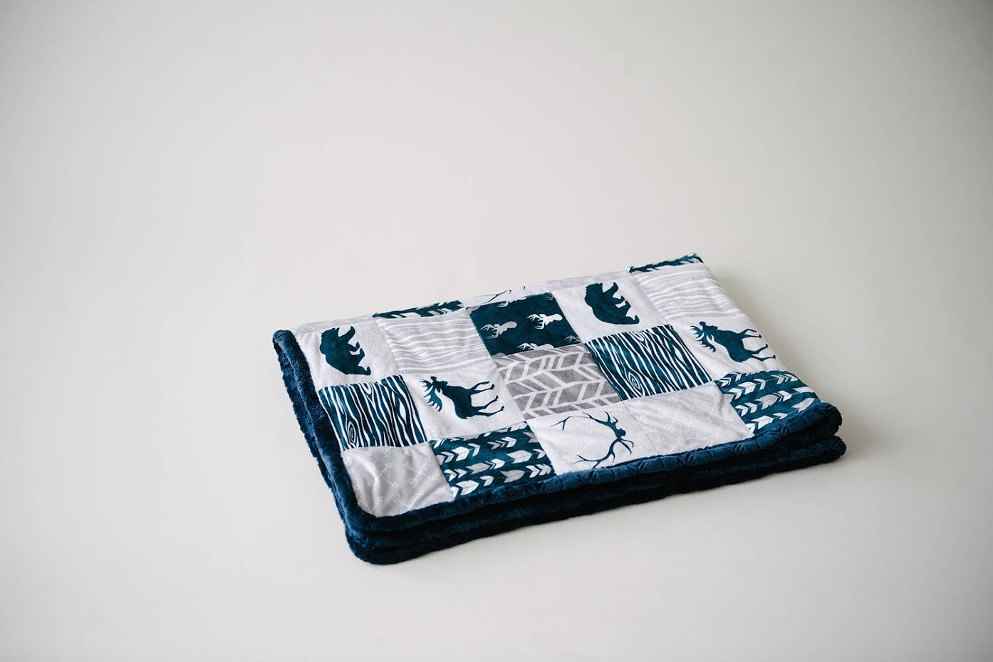 May include: A folded patchwork blanket featuring a variety of patterns and animal silhouettes in teal and gray on a white background. The blanket has a plush, dark blue border. The design includes bears, moose, and abstract patterns.