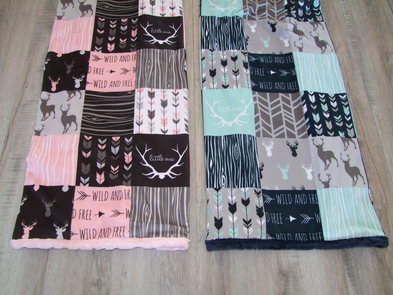 May include: Two patchwork blankets with deer, arrow, and wood grain patterns. One blanket has pink and black squares with the text "WILD AND FREE" and "little one". The other has teal and gray squares.