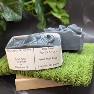 May include: Two gray, rectangular bars of Tea Tree Facial Soap made with goat milk, resting on a green washcloth. The soap is labeled "Ridgedale Farms" and "Small Batch Handmade." Each bar weighs 4 ounces (113 grams).
