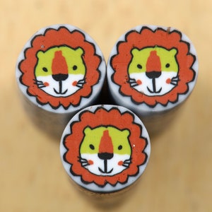 May include: Three round polymer clay beads with a cartoon lion face design. The lion faces are yellow, green, and orange with white accents. The beads are on a light brown surface.