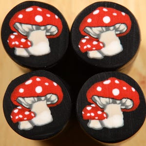May include: Four circular, black wooden plugs featuring a red mushroom design. Each mushroom has a red cap with white spots, a white stem, and a smaller mushroom. The plugs are arranged in a square formation.