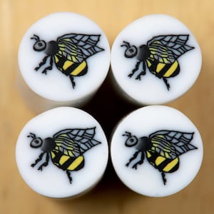 Puede incluir: Cuatro azulejos redondos de cerámica blanca con ilustraciones de abejas negras y amarillas. Cada azulejo presenta una abeja con las alas extendidas y un cuerpo rayado negro y amarillo.