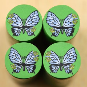 Puede incluir: Cuatro posavasos redondos verdes con un diseño de mariposa azul y negra. Cada mariposa tiene flores amarillas en sus alas.