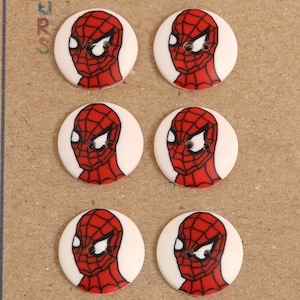 May include: Six round white buttons with a red and black Spiderman image on each. The buttons have a white background and a black outline.
