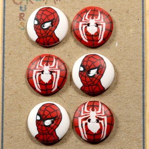 Peut inclure: Six boutons ronds avec une base blanche et des motifs Spider-Man rouges et noirs. Trois boutons présentent le masque de Spider-Man et trois l'emblème de l'araignée. Les boutons sont sur une surface brun clair.